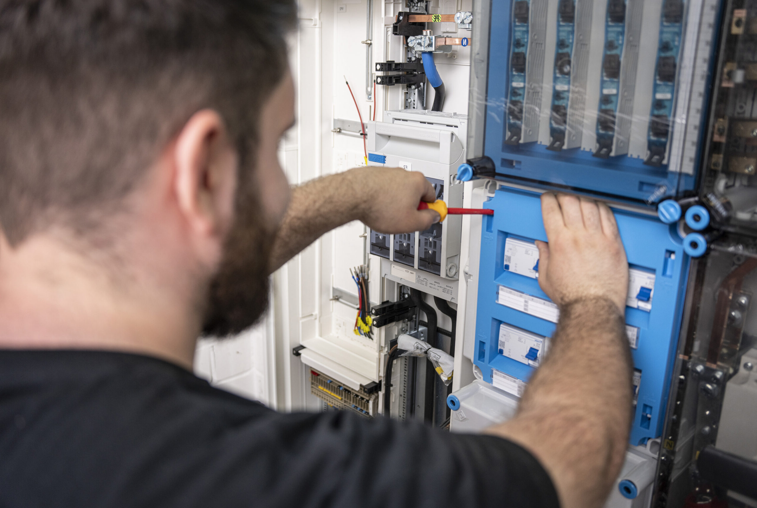 Eine Person arbeitet mit einem Schraubendreher an einem geöffneten Elektroschaltschrank. Blaue und graue Module, Kabel sowie Sicherungen sind sichtbar, die Person ist von hinten zu sehen.