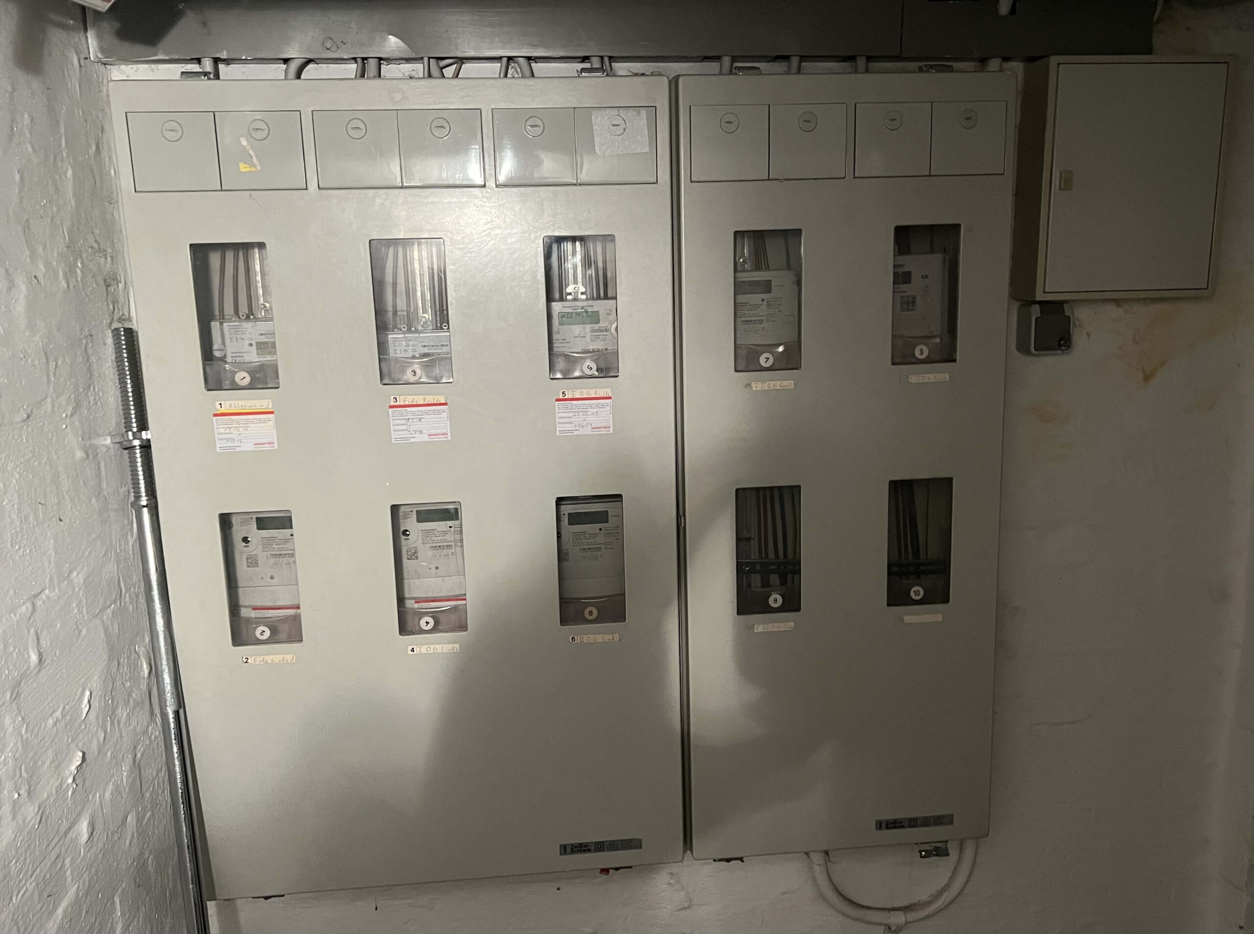 Eine ältere Zählertafel mit einem digitalen Stromzähler. Darüber sind zwei Sicherungskästen von Hager und einem weiteren Hersteller montiert. Die Wand ist weiß gestrichen, und Kabel verlaufen entlang der Decke.