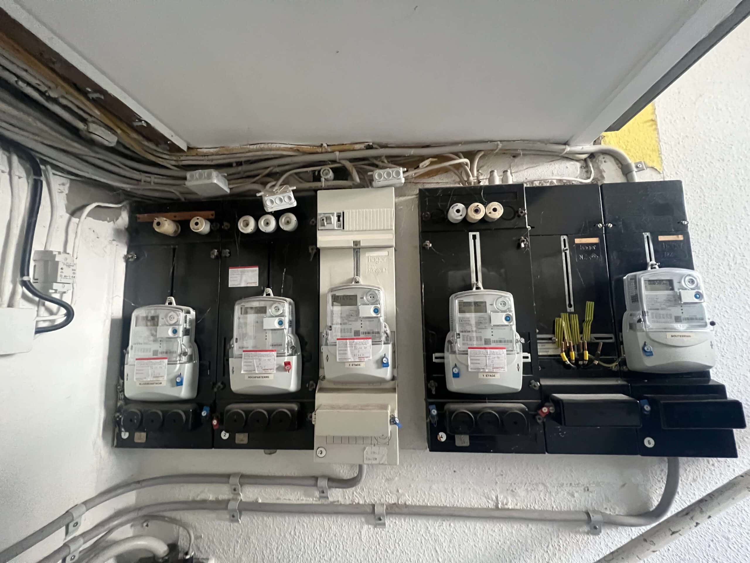 Eine alte Zählertafel mit zwei digitalen Stromzählern und einem zusätzlichen kleinen Zähler in der Mitte. Die Schalttafel ist schwarz mit weißen Schraubsicherungen und einer modernen Sicherungseinheit rechts.