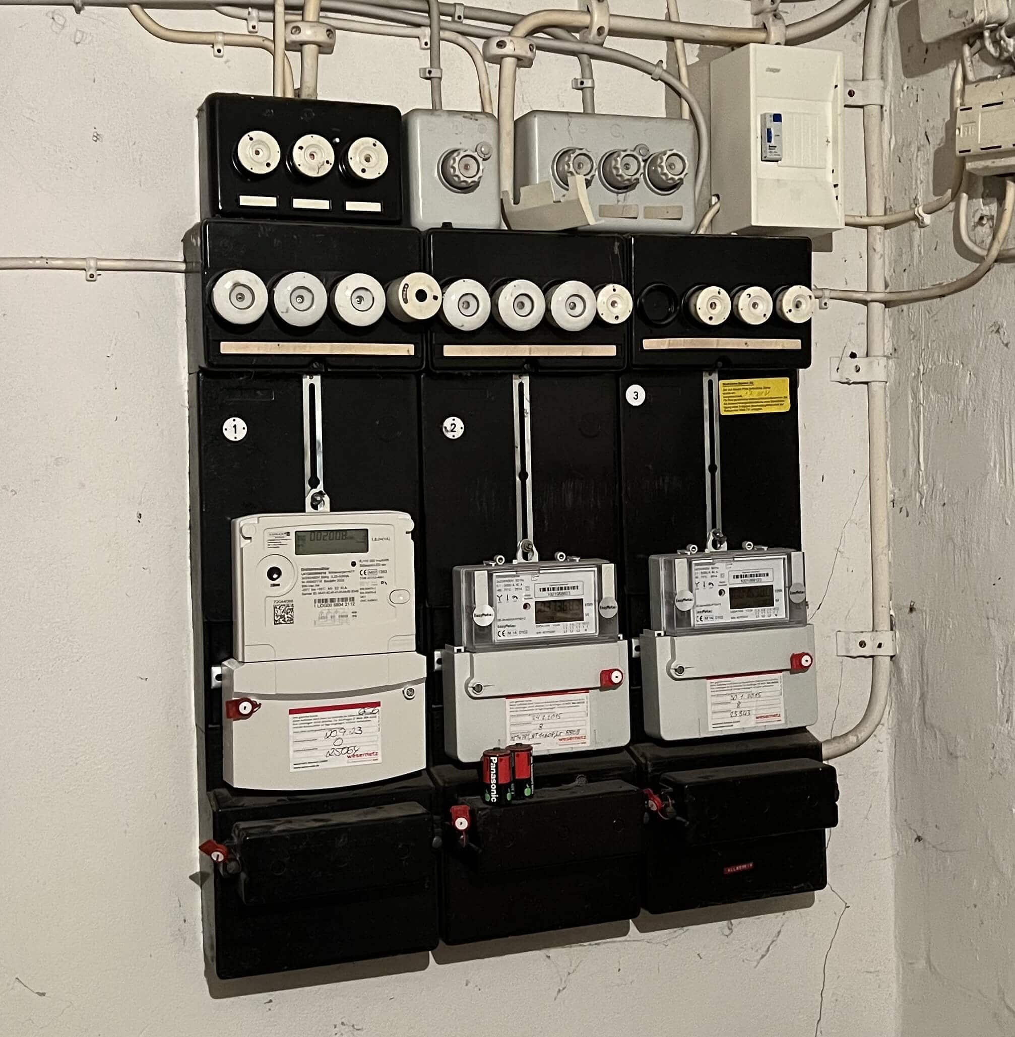 Eine alte Zählertafel mit vier digitalen Stromzählern, montiert auf schwarzen Schaltschränken. Oben sind mehrere Schraubsicherungen zu sehen, während zahlreiche Kabel an der Decke entlang verlaufen.