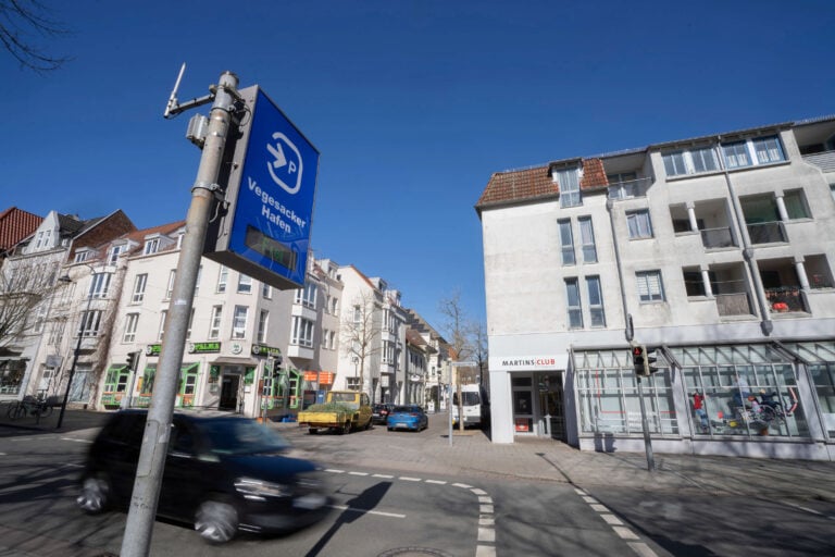 Straßenszene in Bremen-Vegesack mit einem blauen Hinweisschild für den Parkplatz ‚Vegesacker Hafen‘. Im Hintergrund befinden sich Geschäfte und Wohnhäuser.