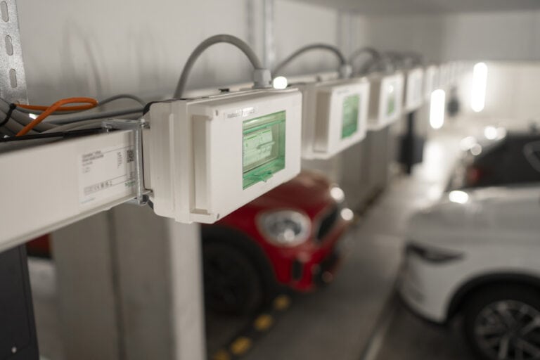 Reihe von Verteilerkästen an der Decke einer Tiefgarage, im Hintergrund sind mehrere geparkte Fahrzeuge sichtbar.