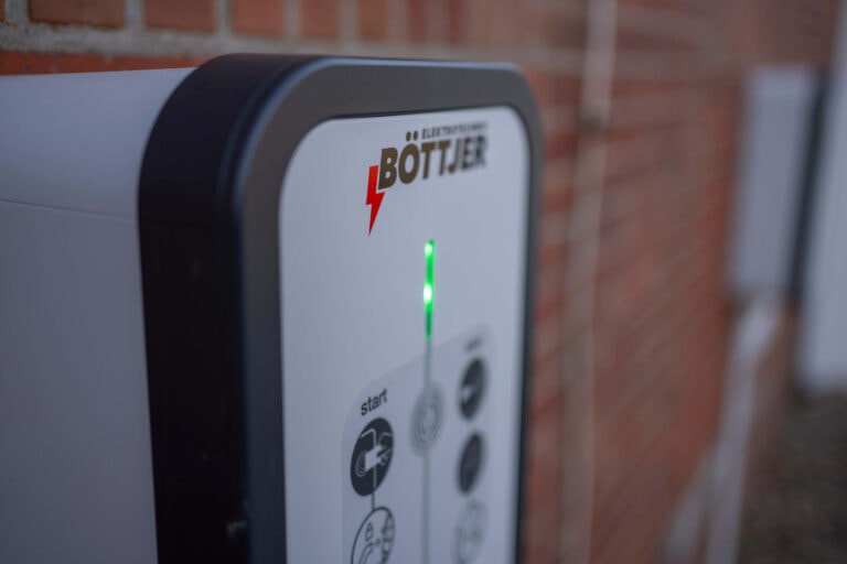 Nahaufnahme einer weißen Ladestation mit der Aufschrift „ELEKTROTECHNIK BÖTTJER“ und einem roten Blitzsymbol. Eine grüne LED-Leuchte ist in der Mitte der Station aktiv. Die Beschriftung und Piktogramme weisen auf Bedienfunktionen wie Start und Stop hin.