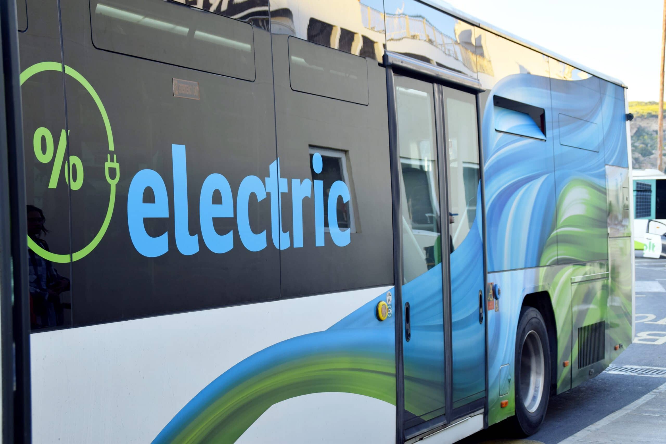 Seitenansicht eines weiß-blauen Elektrobusses mit großer, auffälliger Beschriftung „electric“ in blauer Schrift auf schwarzem Hintergrund. Links neben dem Text ist ein grünes Symbol in Form eines Steckers mit Prozentzeichen zu sehen. Der Bus ist mit einem großflächigen, wellenförmigen Muster in Blau- und Grüntönen gestaltet.