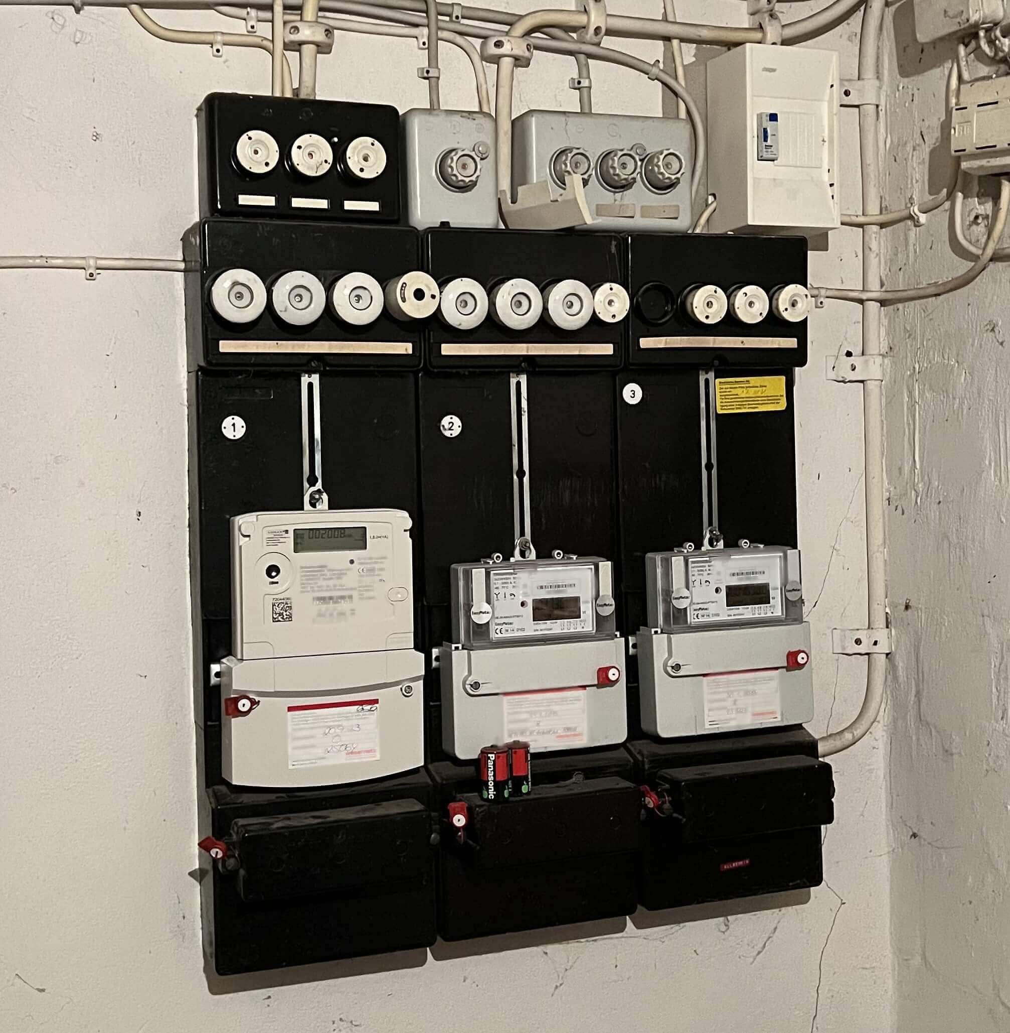 Eine alte Zählertafel mit vier digitalen Stromzählern, montiert auf schwarzen Schaltschränken. Oben sind mehrere Schraubsicherungen zu sehen, während zahlreiche Kabel an der Decke entlang verlaufen.