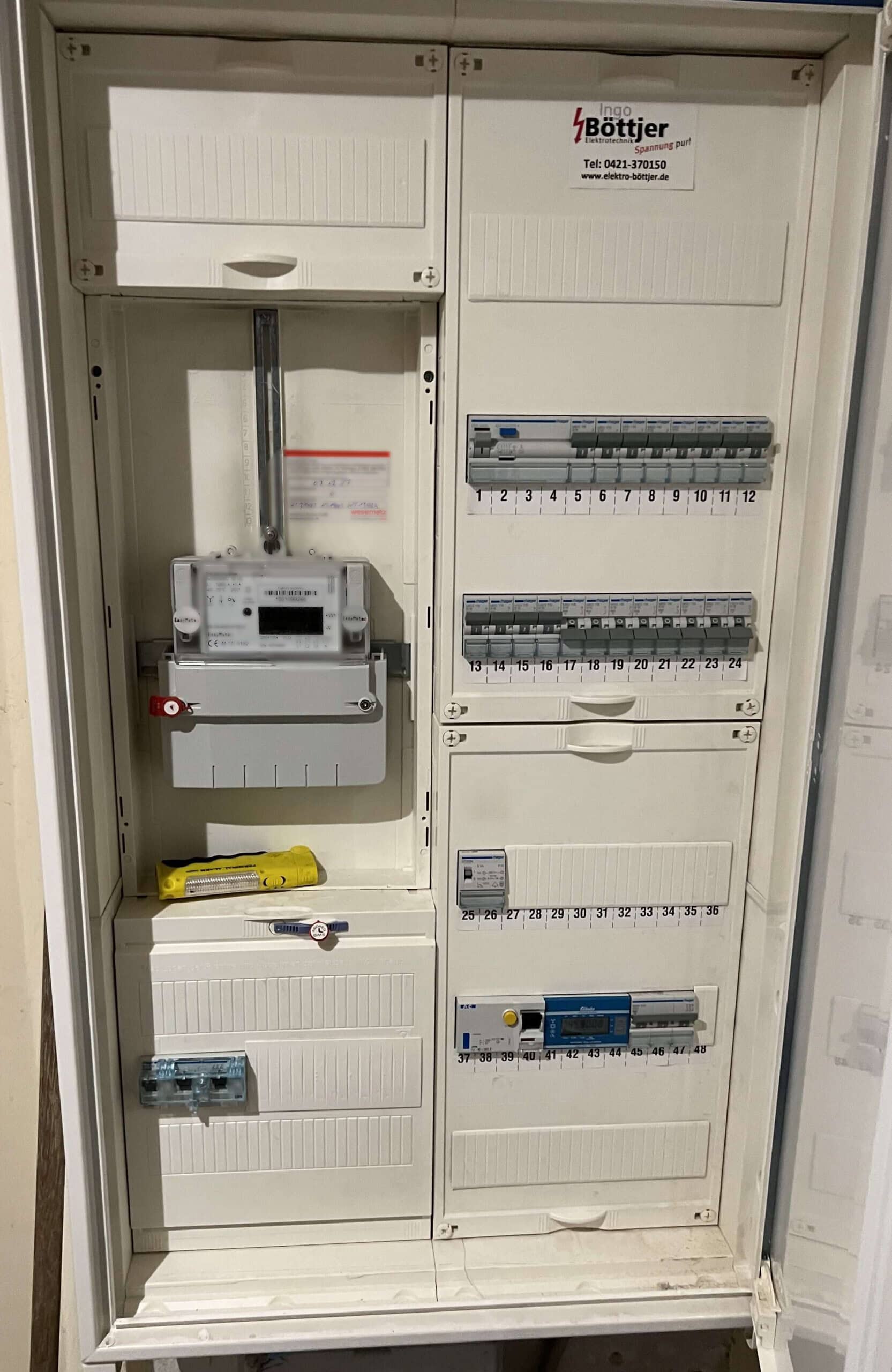 Geöffneter Schaltschrank mit einem digitalen Stromzähler und mehreren Sicherungsreihen. Das Gehäuse ist weiß, mit einem gelben Prüfgerät, das unten im Schrank liegt. Auf der oberen rechten Seite befindet sich ein Aufkleber mit der Aufschrift „Ingo Böttjer – Elektrotechnik“.