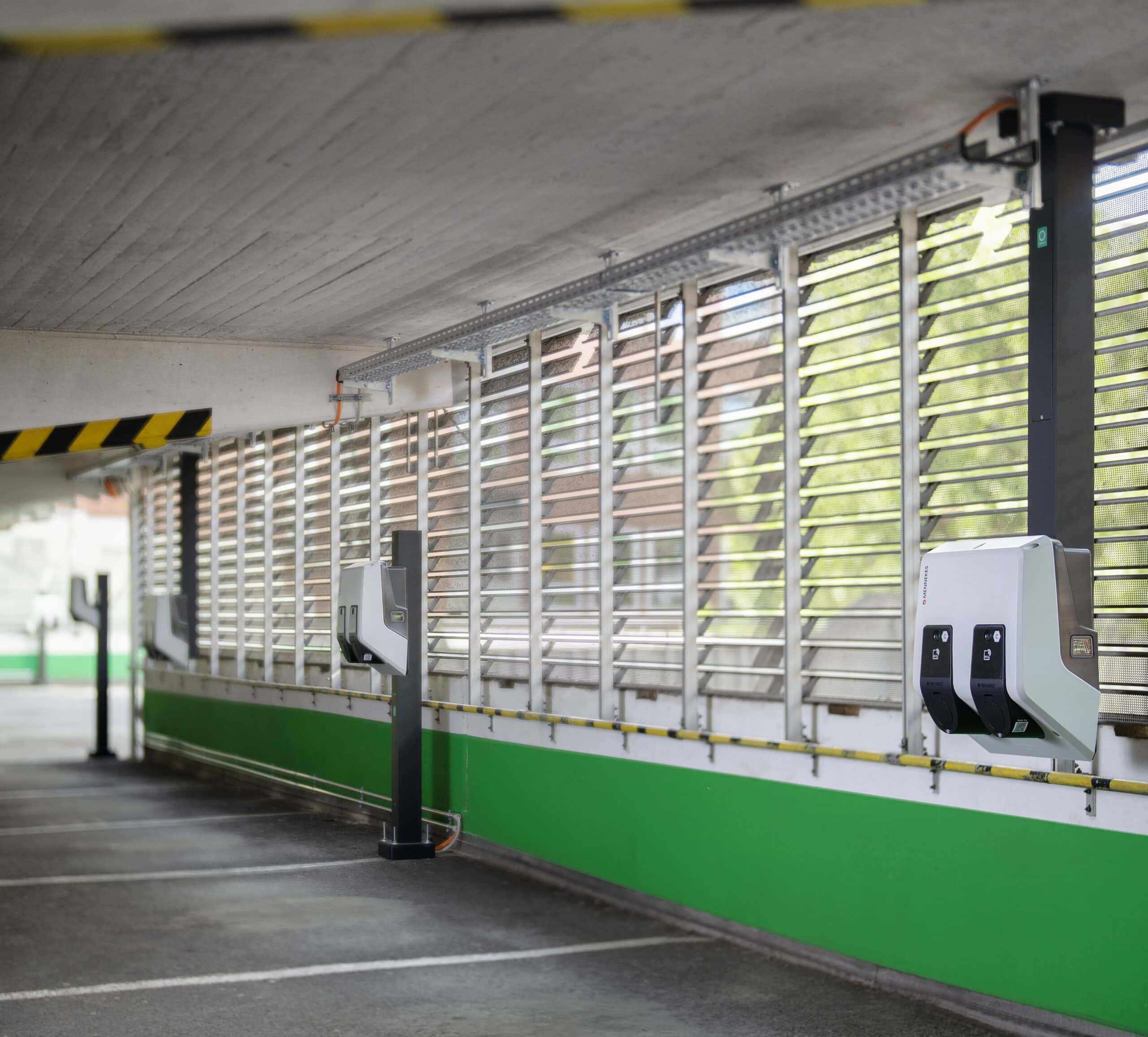 Überdachte Parkgarage mit mehreren leeren Stellplätzen. An der rechten Wand sind zwei weiße E-Ladestationen montiert, davor Lamellenfenster mit Tageslicht und grün-weißer Wandgestaltung.